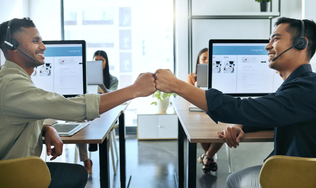 Two diverse male salespeople giving each other a fist bump at their desks.