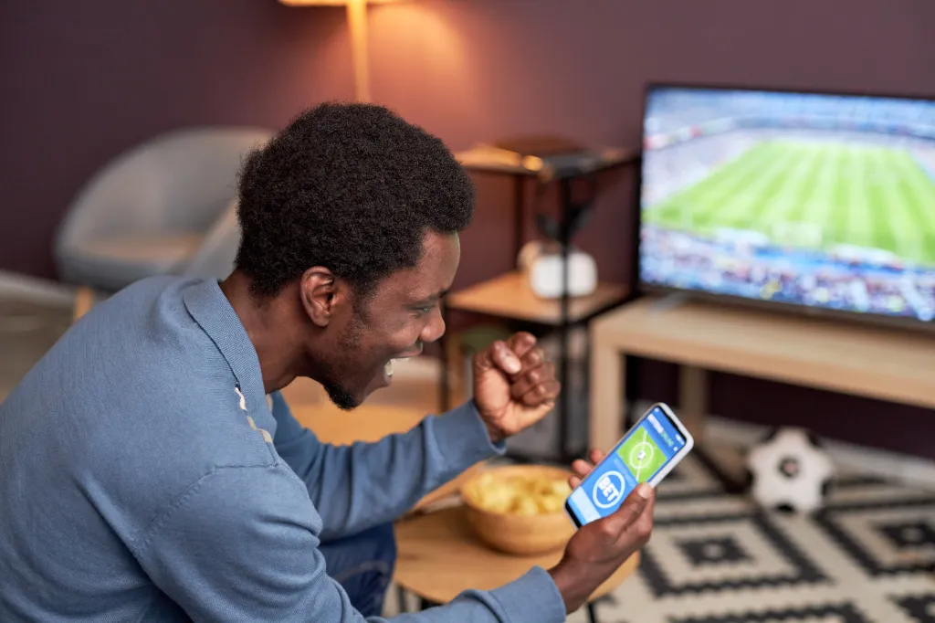 Man holding a smartphone with an online sports betting app and cheering while watching a football match.