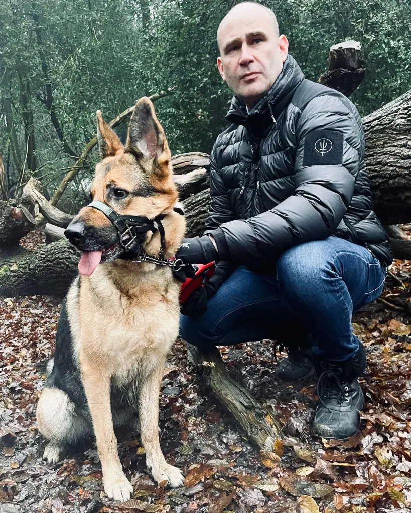 A man kneeling next to a German Shepherd dog in a wooded area.