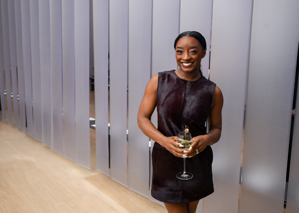 Simone Biles smiling while holding a drink in a dark brown dress.
