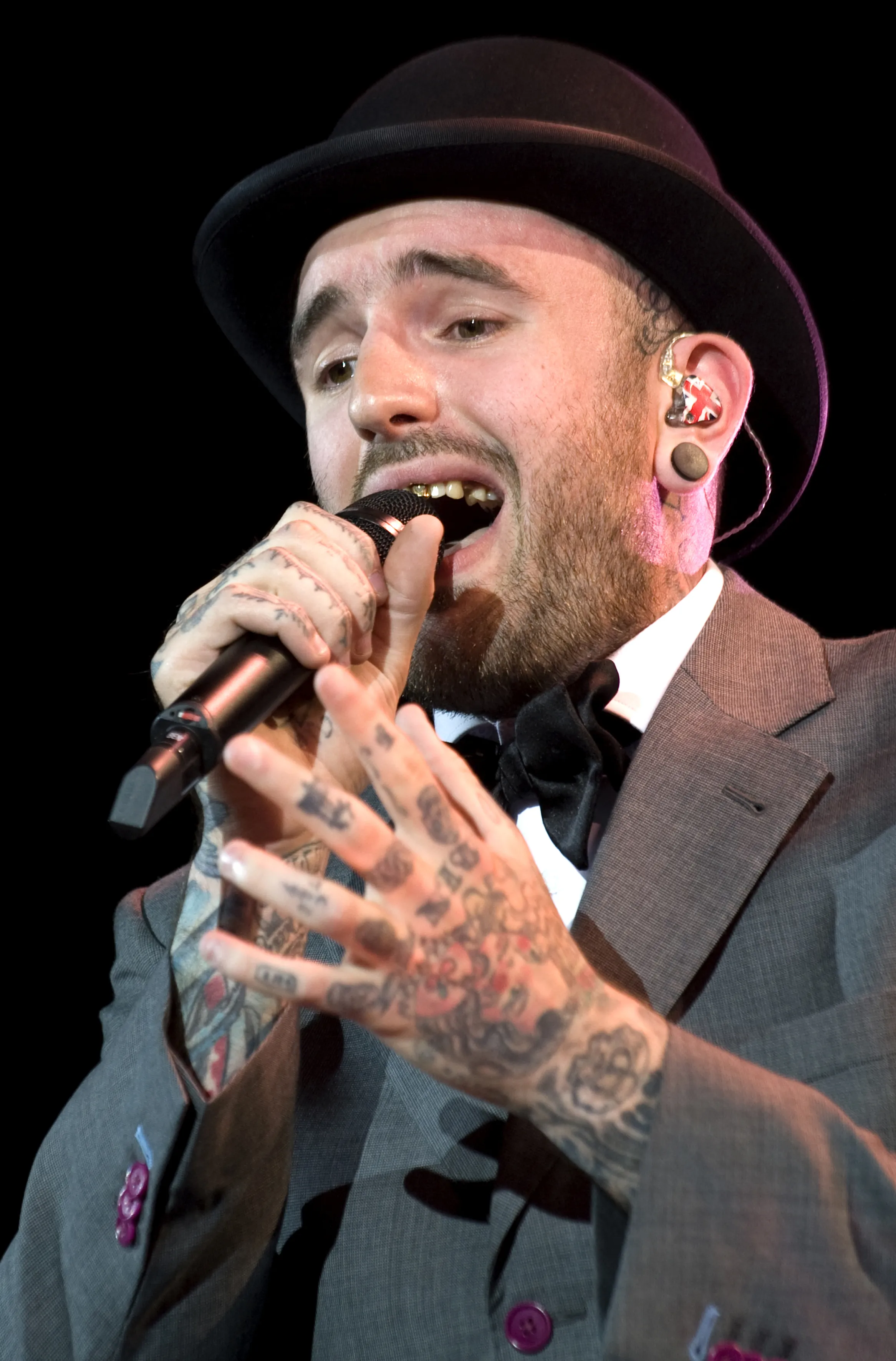 Ben Saunders performing live, wearing a bowler hat, grey suit, bow tie, and holding a microphone.
