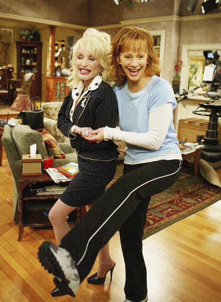 Dolly Parton and Reba McEntire pose together, holding hands, with McEntire holding one leg up as if dancing, on the set of the TV show