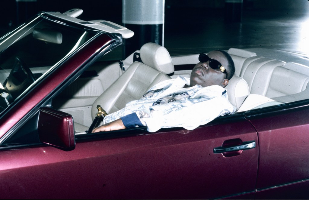 Notorious B.I.G. reclines in a convertible with a cane, wearing sunglasses and a patterned shirt, on the set of the 