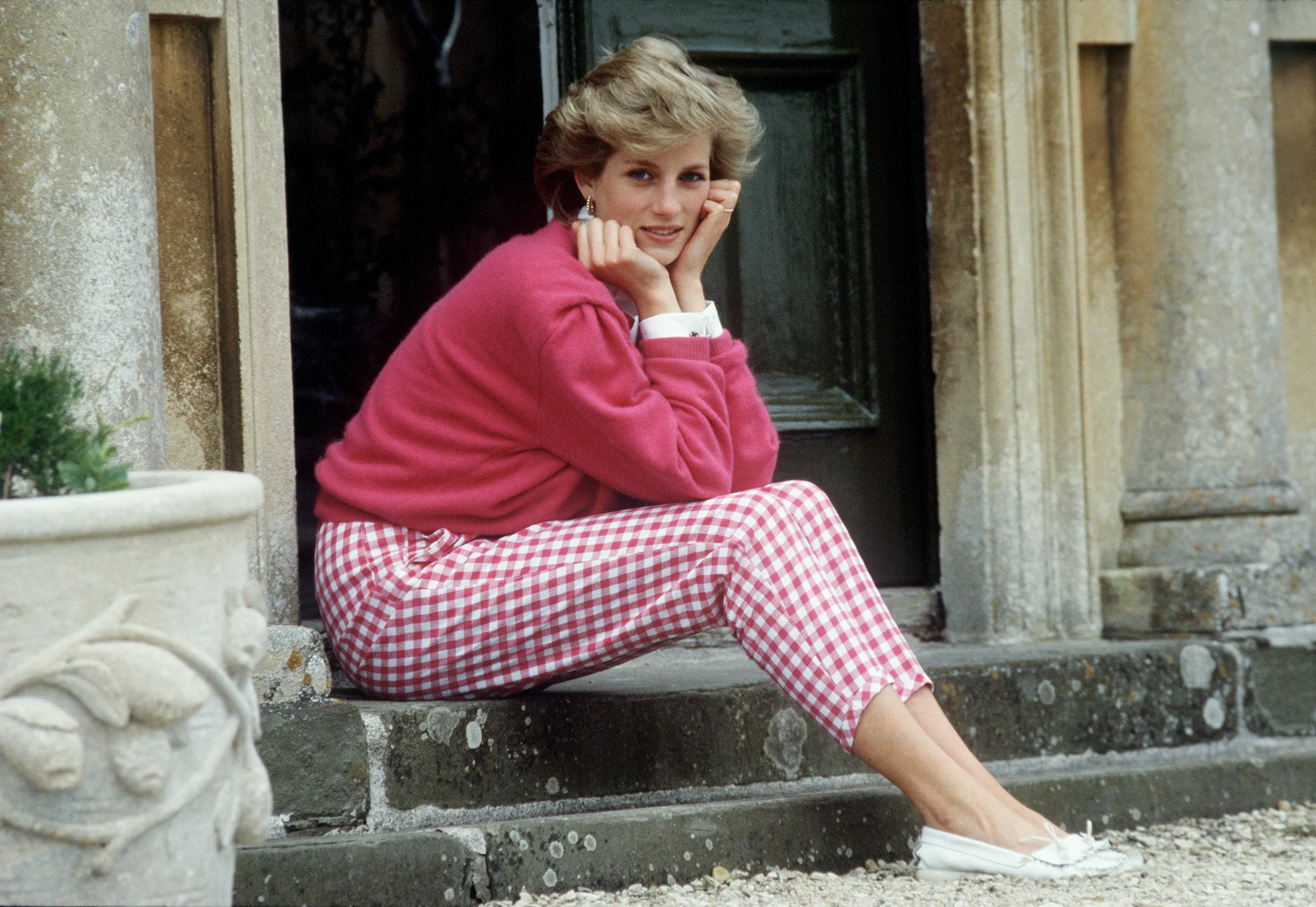 Princess Diana sitting on a stone step, wearing a pink sweater and red and white gingham pants, with her hands resting under her chin.