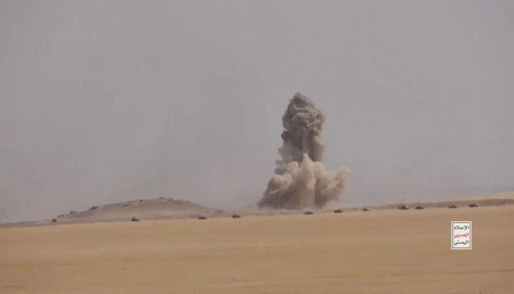 Smoke from an explosion billows into the sky above a desert landscape with military vehicles, depicting a battle between Iran-aligned Houthi fighters and Saudi-backed forces in al Jawf governate.