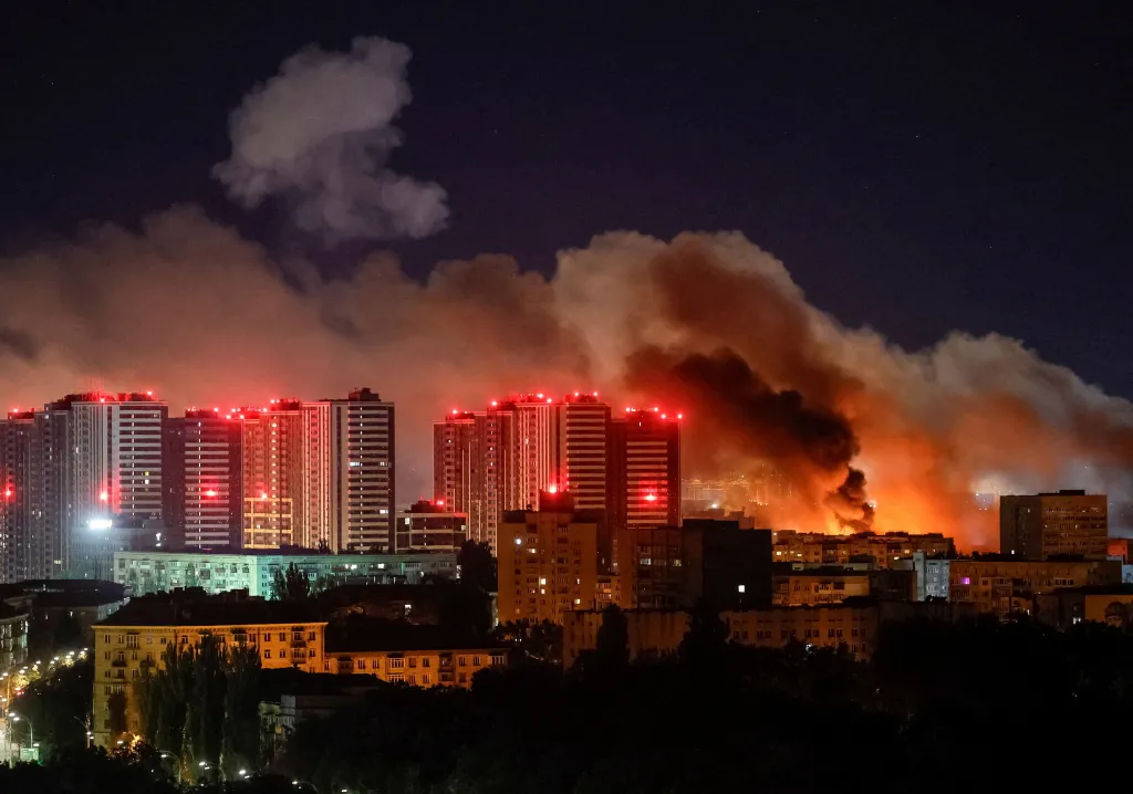 Smoke rises from a Russian missile strike in Kyiv at night, with multiple high-rise buildings illuminated by red lights in the foreground.