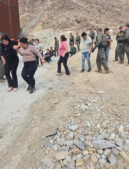 Law enforcement officials detaining a group of people near a border wall in a desert environment.