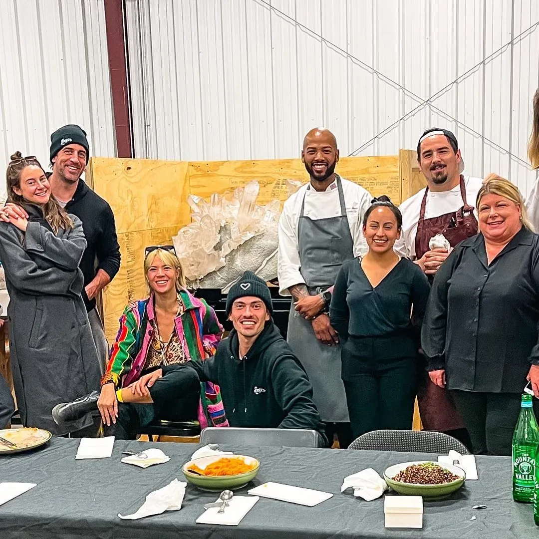 Aaron Rodgers and Shailene Woodley holding hands at a dinner table with others, including chefs.