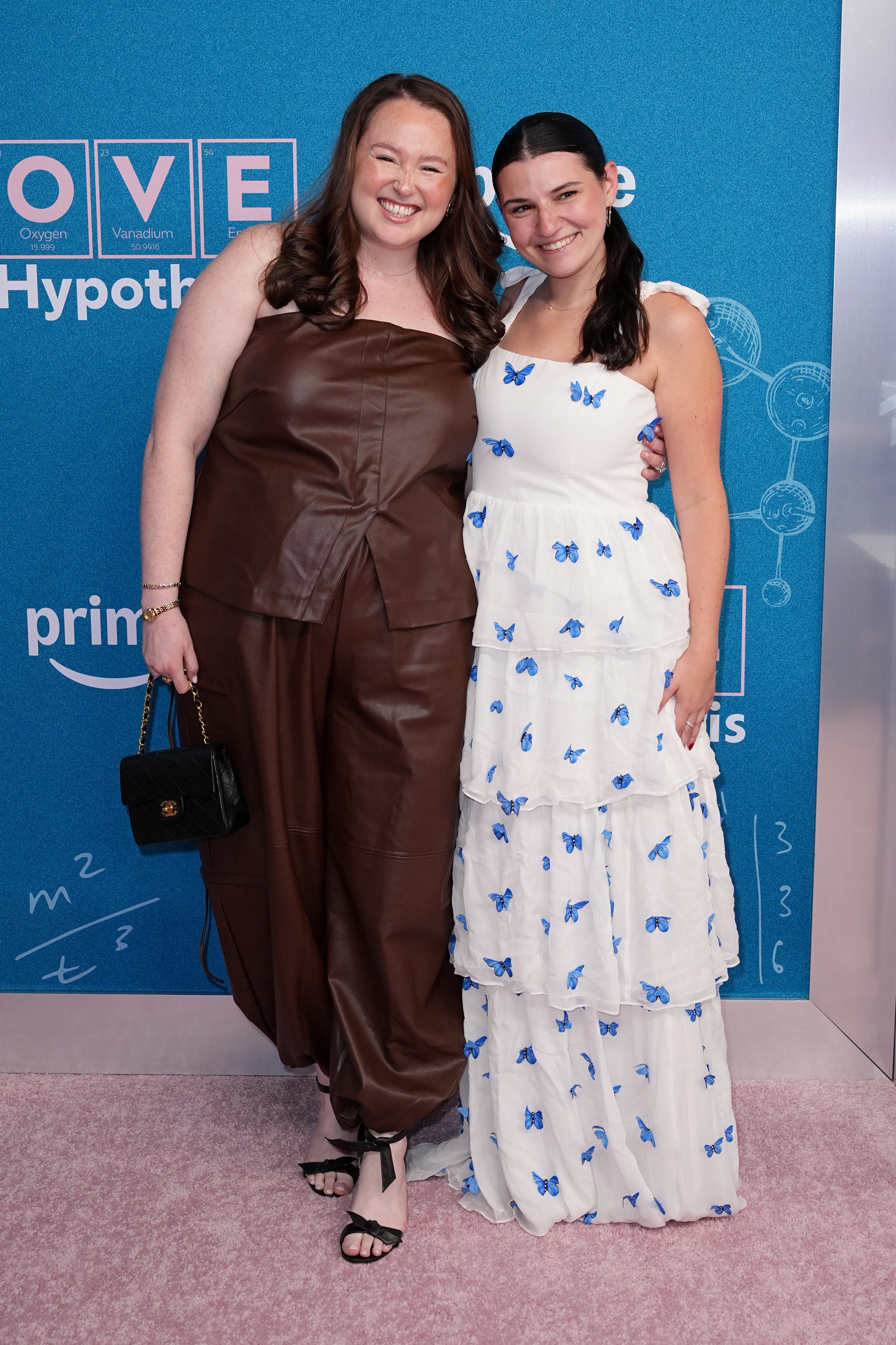 Sophie Beren in a brown leather outfit and Carly Heitner in a white dress with blue butterflies pose at 