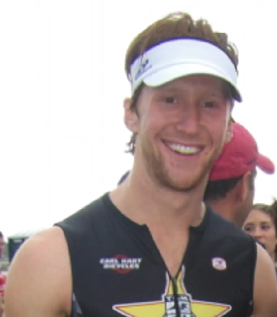 Spencer Korwin, former Team USA triathlete, smiling and wearing a white visor and a black sleeveless shirt with a yellow star design.