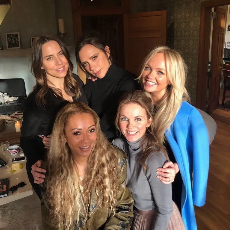 The Spice Girls members Mel B, Victoria Beckham, Geri Halliwell, Emma Bunton, and Mel C smiling together in a reunion photo.