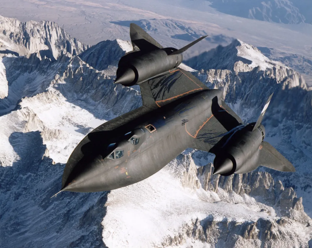 An SR-71B Blackbird aerial reconnaissance aircraft flying over snow-capped mountains.
