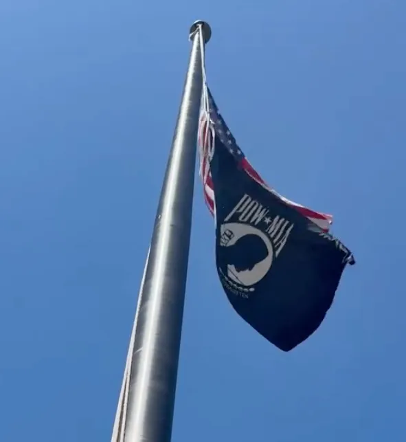 A POW/MIA flag and an American flag flying high on a flagpole against a blue sky.