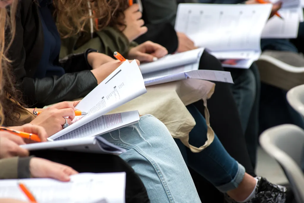 Students writing a test with paper questionnaires and orange pens.