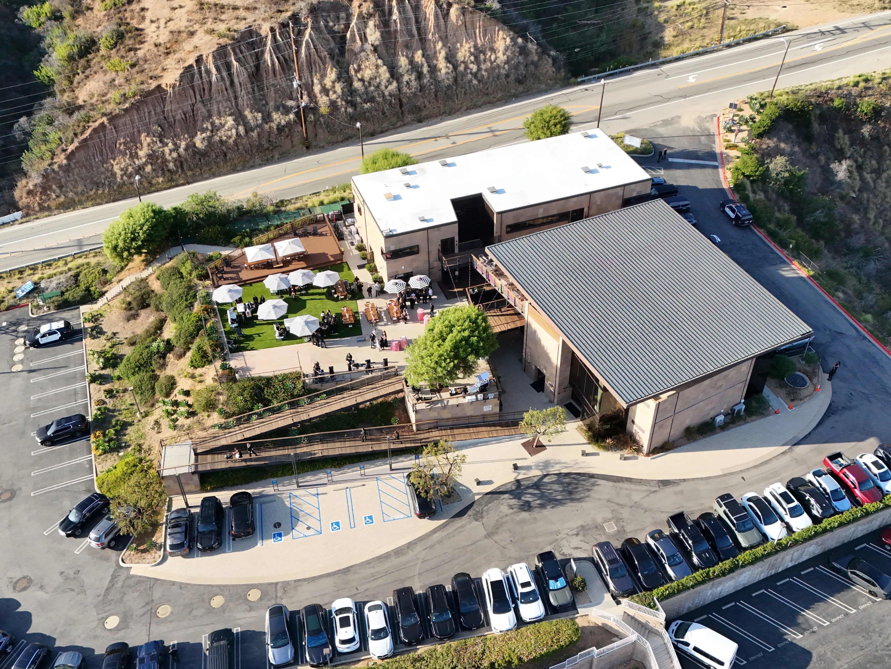 Aerial view of Malibu Pacific Church, a large building with a white roof and outdoor patio with umbrellas, surrounded by a parking lot and a road.