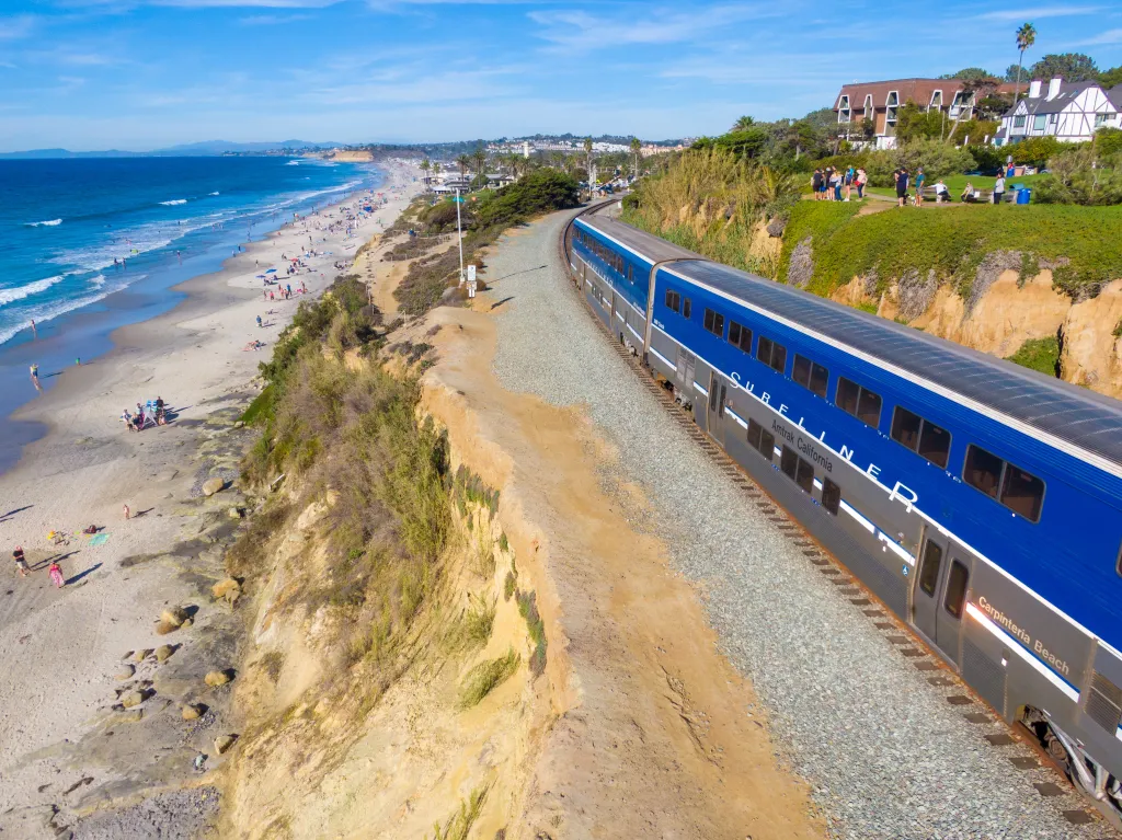 Drone image of a southbound Amtrak Surfliner train traveling along the coast in Del Mar, California, with beachgoers on the sand.