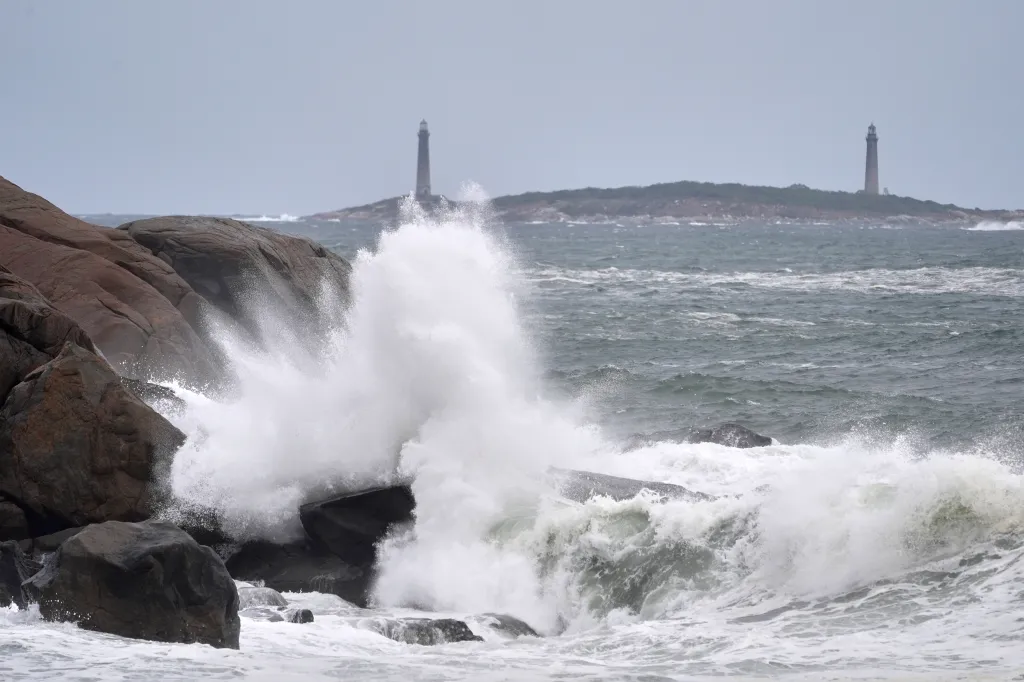 Waves crash against a rocky coastline with two lighthouses in the distance.
