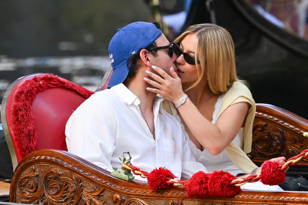 Sydney Sweeney, in a white dress and yellow sweater, and Scooter Braun, in a white shirt and blue baseball cap, share a romantic kiss while enjoying a gondola ride through the canals of Venice.