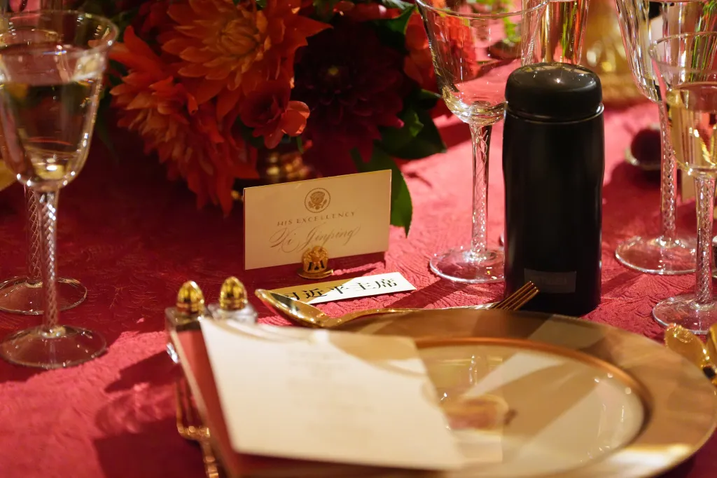 A table setting for Xi Jinping at a State Dinner, including a place card, water bottle, and floral centerpiece, on a red tablecloth.