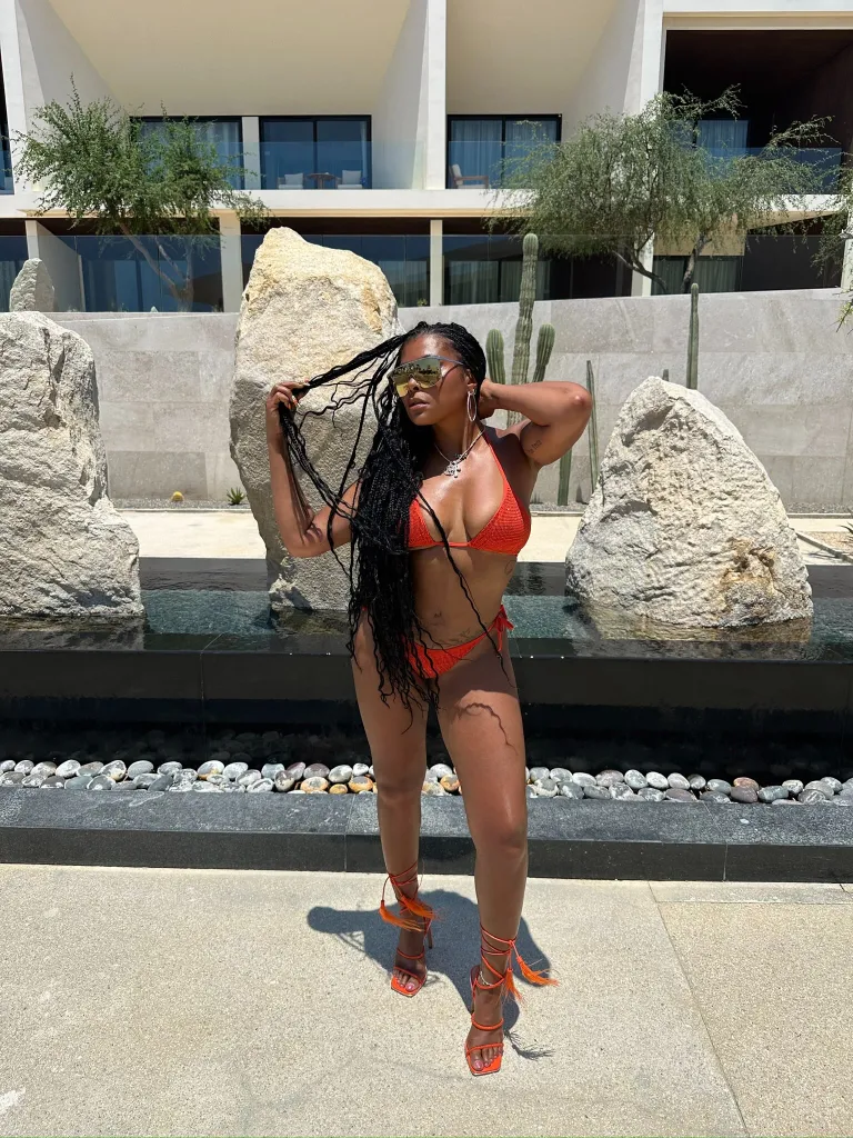 Taraji P. Henson in an orange bikini and heels, posing outdoors next to a pool with large rocks and cacti.