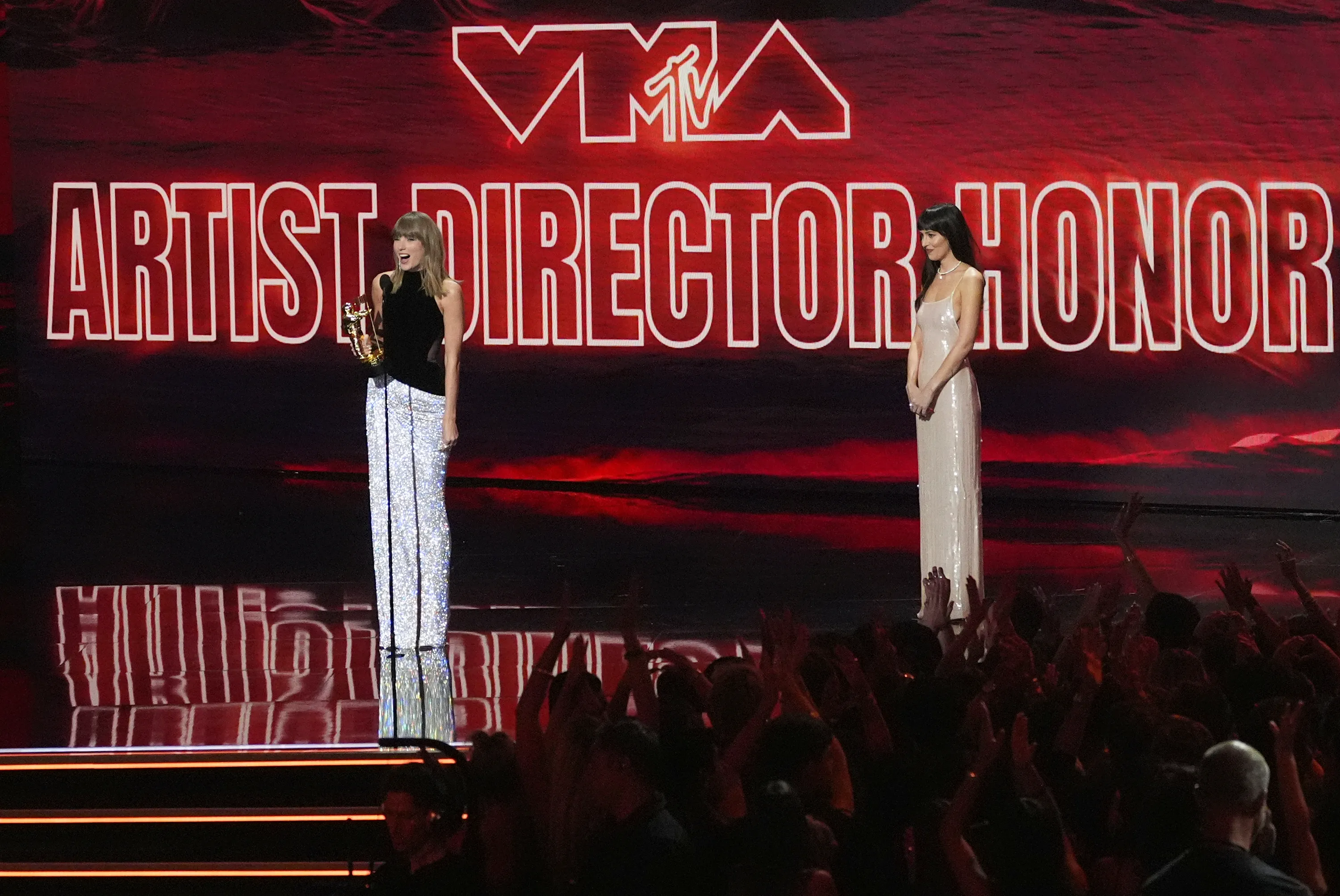 Taylor Swift accepts the MTV VMA Artist Director Honors award while Dakota Johnson looks on, with an excited audience in front of them.