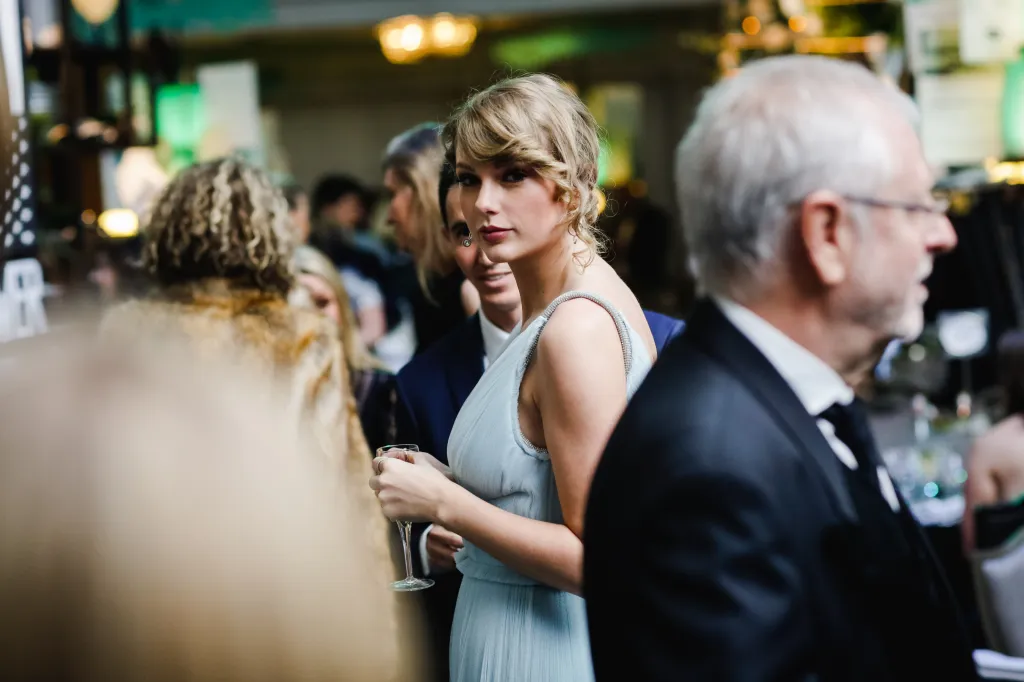 Taylor Swift in a light blue dress looking at the camera, holding a drink, surrounded by people at a party.