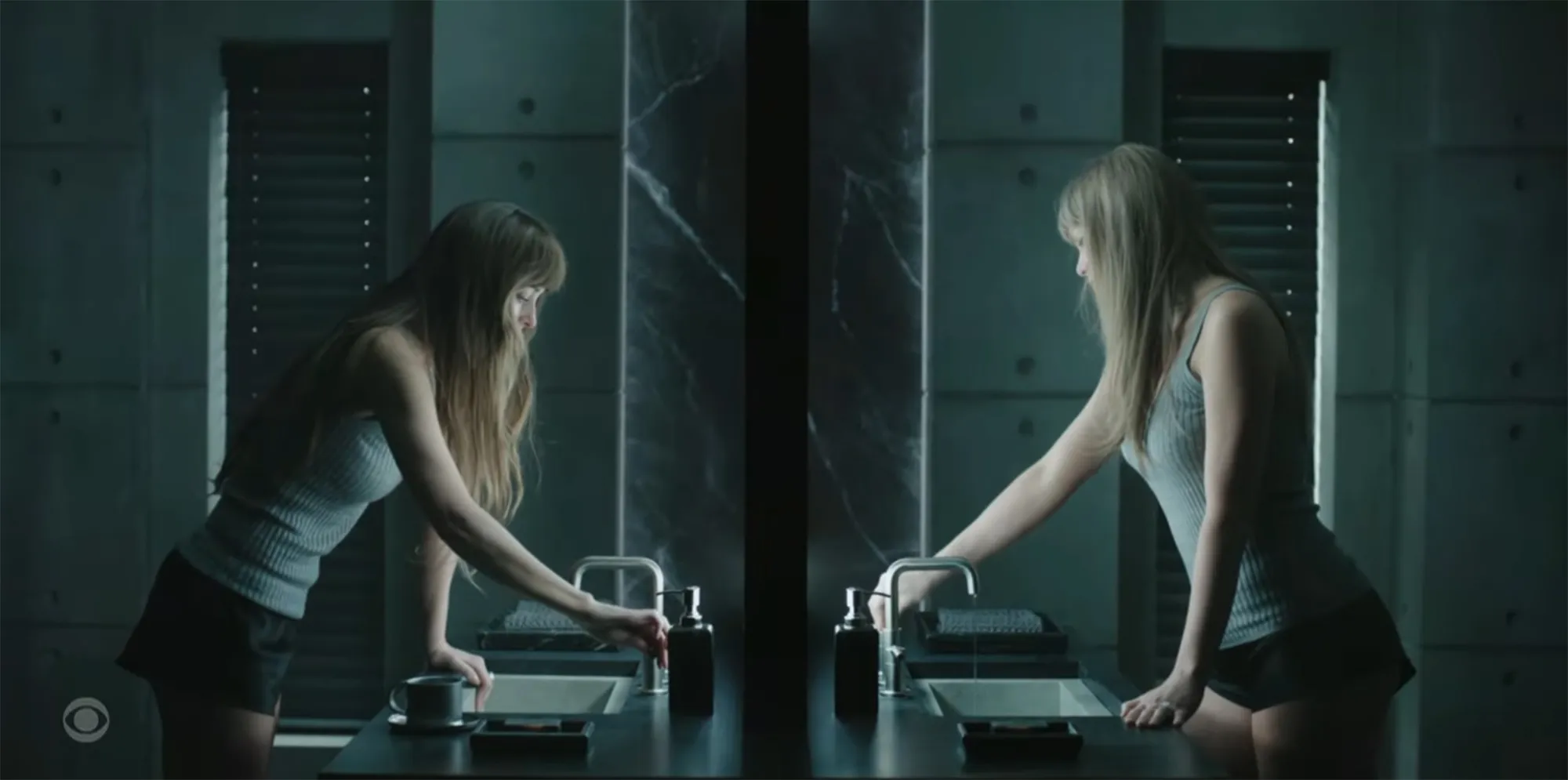 Taylor Swift in a light blue tank top and dark shorts, reaching for a soap dispenser in a dark, modern bathroom with two sinks and a mirror reflecting her.