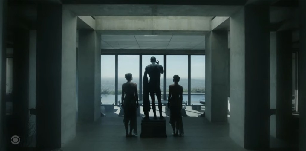 Three figures stand in a modern, angular building, looking out a large window at a distant landscape.