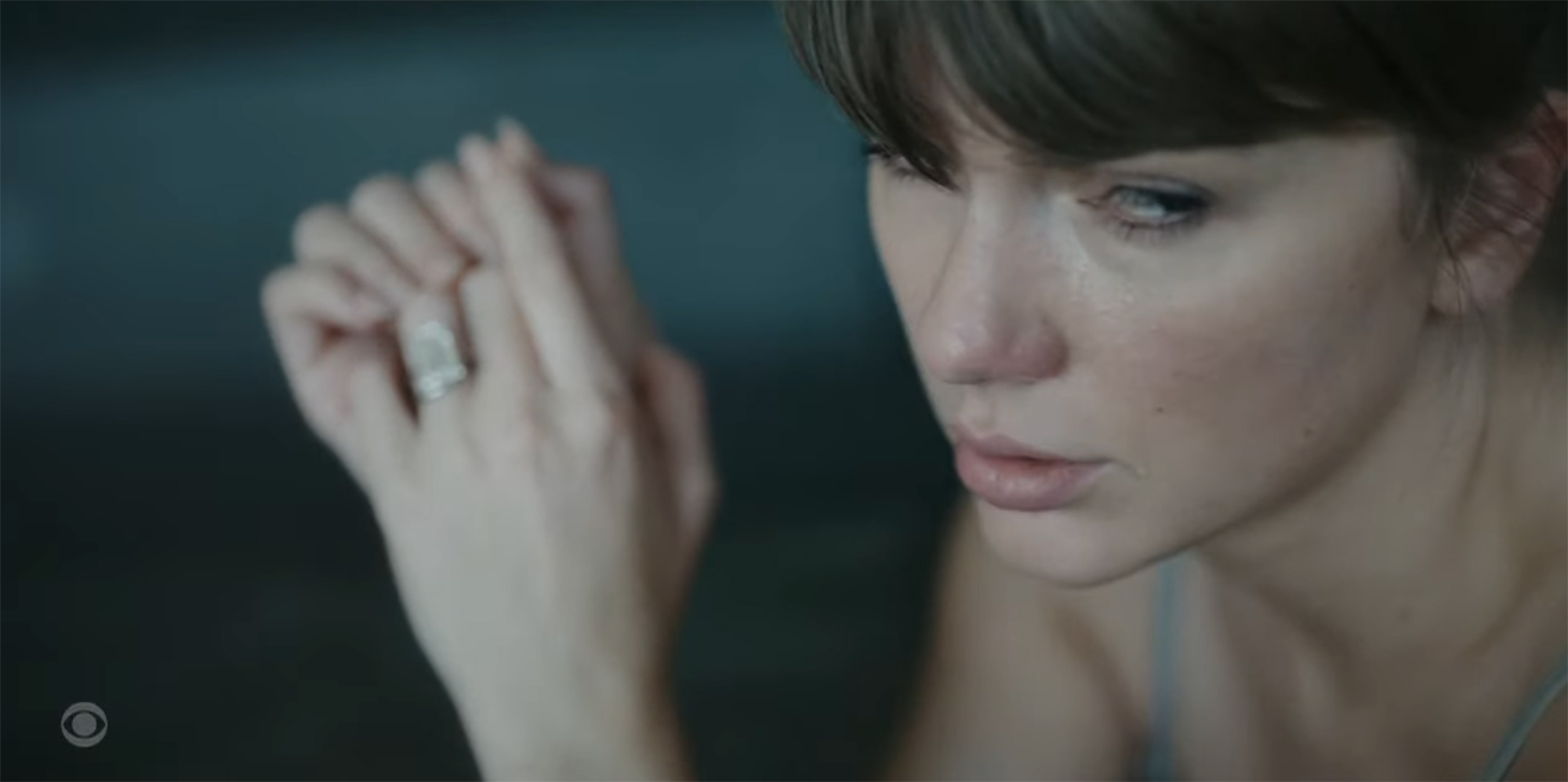 Close-up of Taylor Swift with dark hair and bangs, wearing a silver top, tears in her eyes, and a large diamond ring on her left hand.