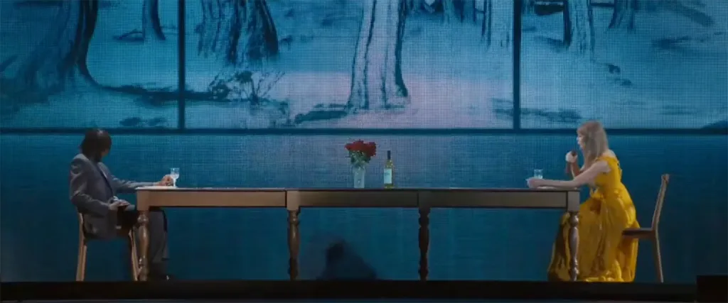 Taylor Swift in a yellow dress sits at a long table across from a man in a gray suit, a vase of red roses and a bottle of wine between them.