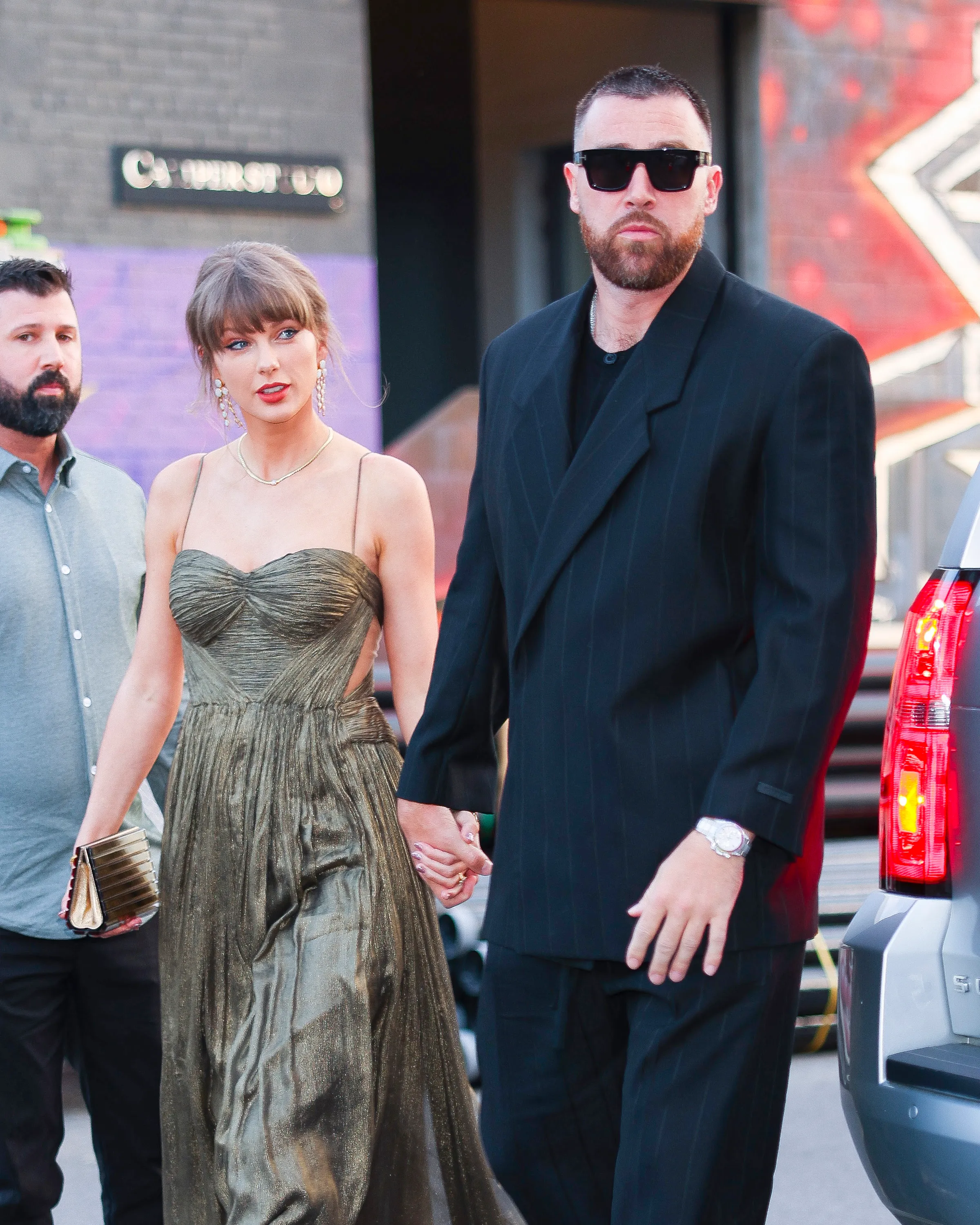 Taylor Swift in a gold dress holding hands with Travis Kelce in a black suit and sunglasses.