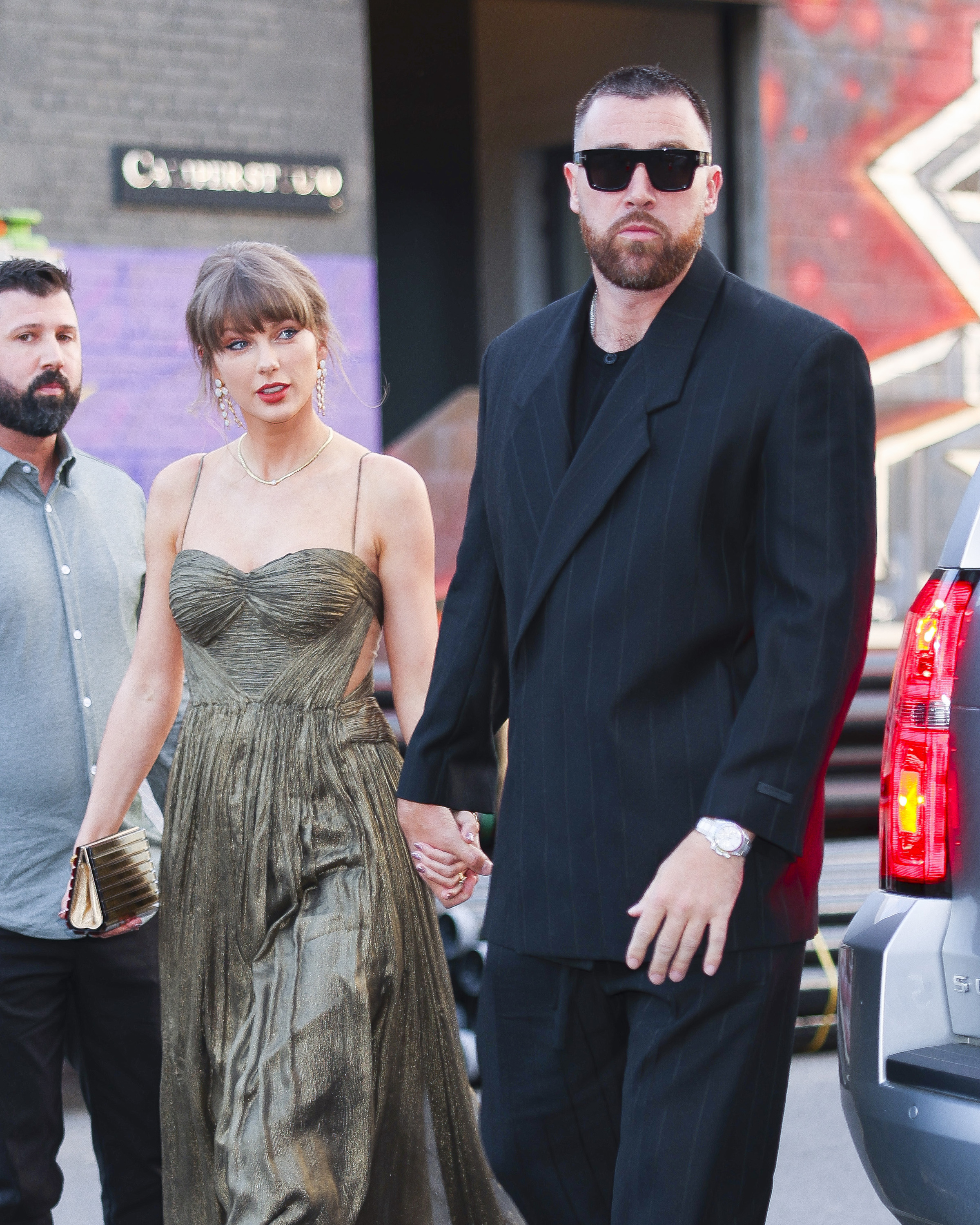 Taylor Swift in a gold gown and Travis Kelce in a dark suit holding hands in Brooklyn.