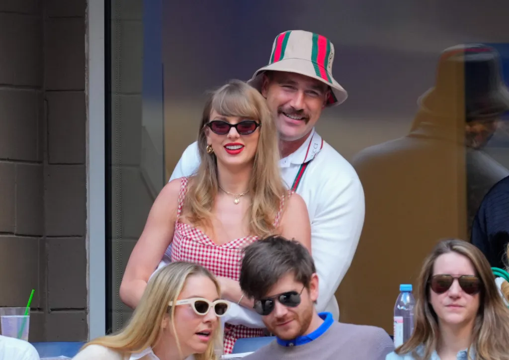 Taylor Swift wearing a red and white checkered dress and red sunglasses, and Travis Kelce wearing a striped bucket hat and white shirt, smiling at the US Open.