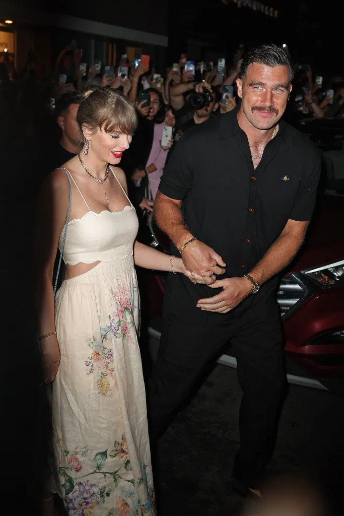 Taylor Swift and Travis Kelce holding hands while walking, with many people in the background taking photos of them.