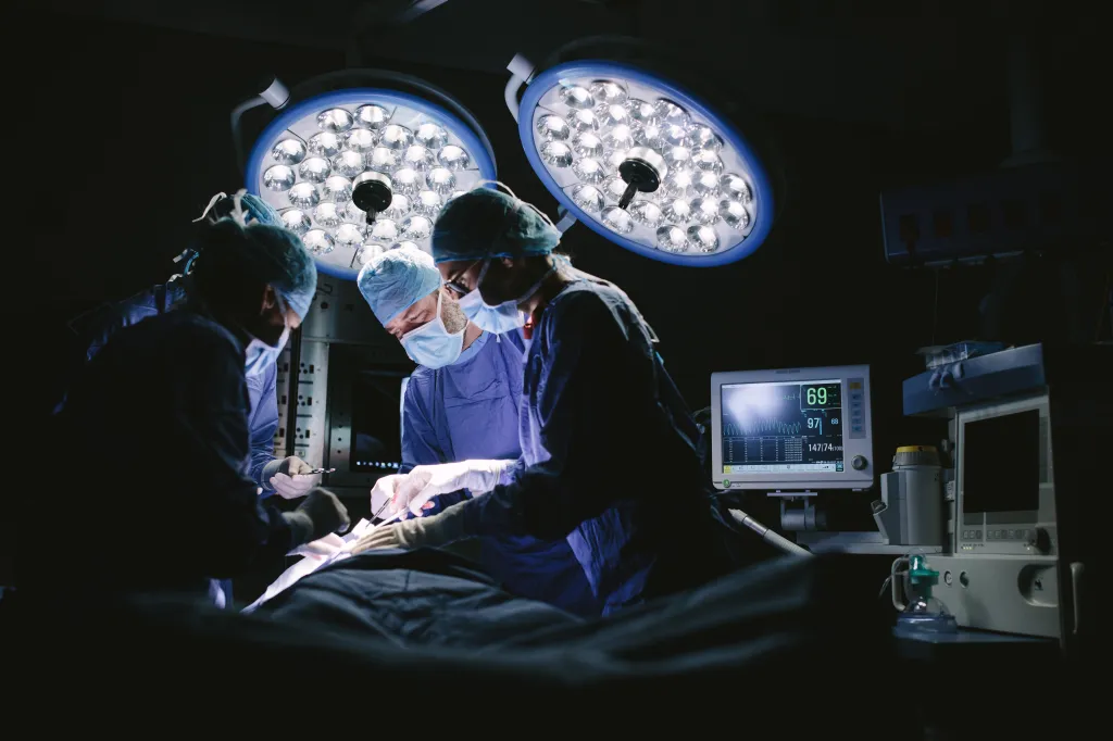 A team of surgeons operating on a patient in an operating room with bright overhead lights and a vital signs monitor.