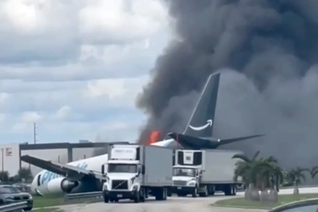 An Amazon cargo jet crashed with smoke billowing from its fuselage, with the tail and right wing visible over two semi-trucks on a road.