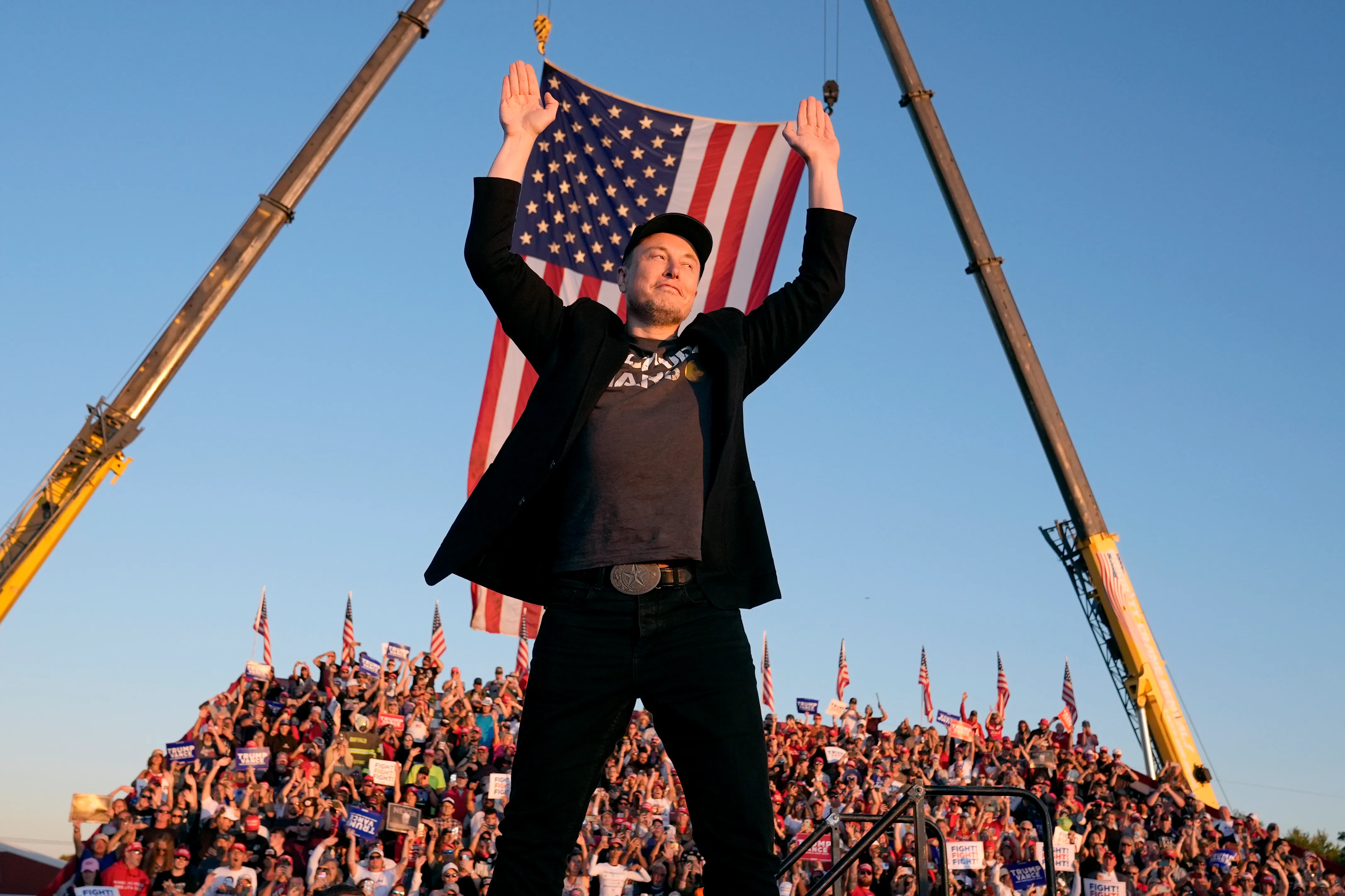Elon Musk stands on a stage with his arms raised, a large American flag behind him, and a crowd of people in front of him.