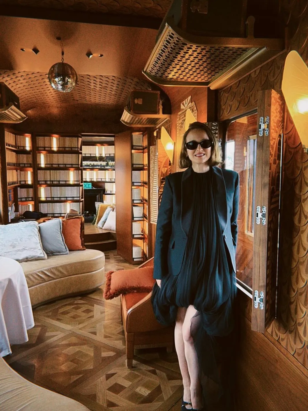 Natalie Portman smiling and posing in a luxurious cabin with dark wood paneling, bookshelves, and an elegant patterned floor.