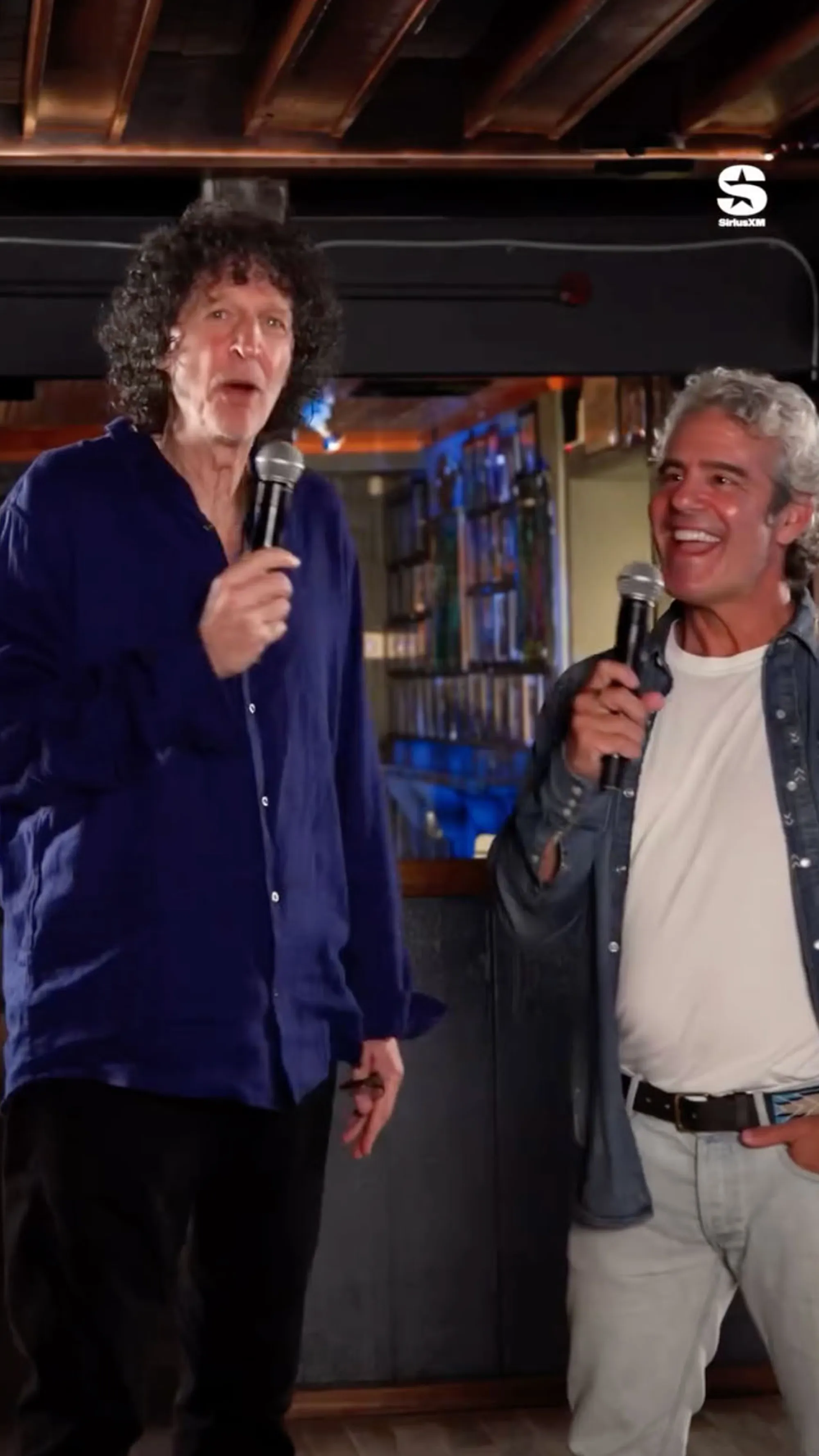 Howard Stern and Andy Cohen holding microphones and smiling.