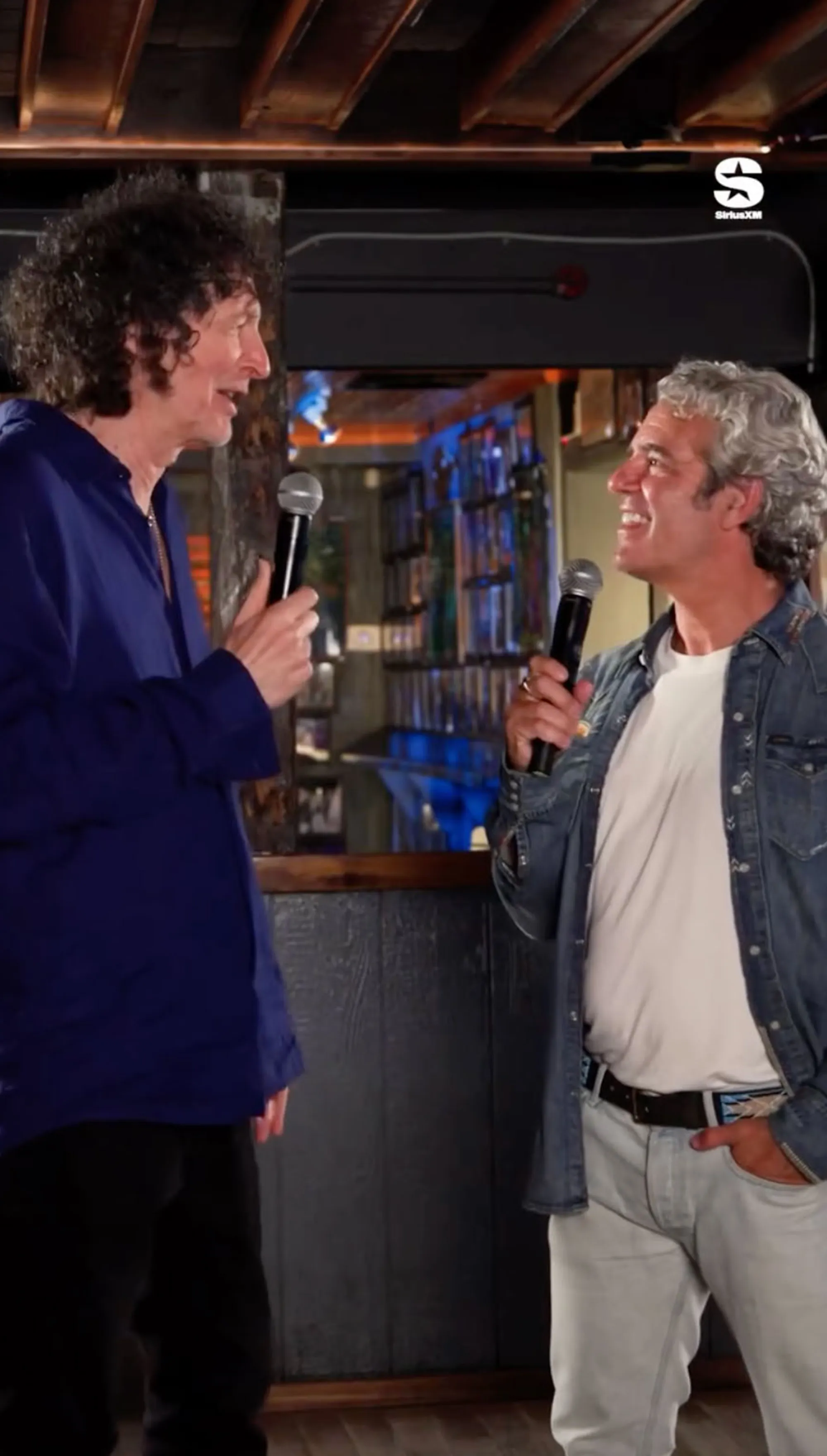 Howard Stern and Andy Cohen holding microphones and smiling at each other.