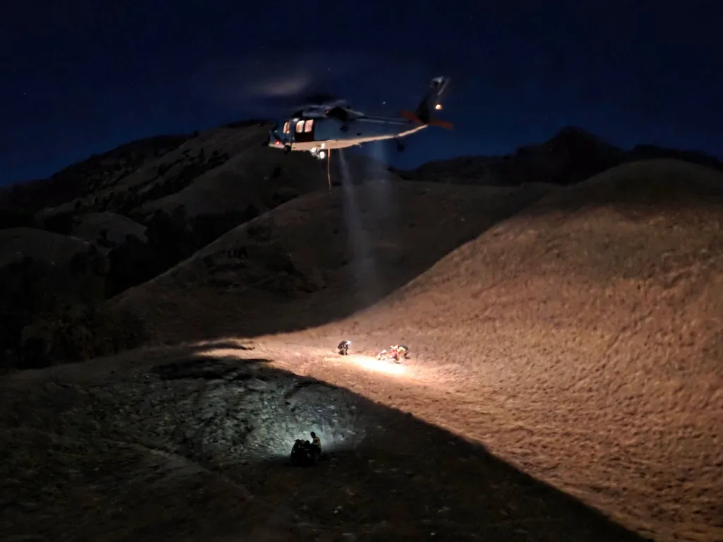 A rescue helicopter illuminates three hikers on a dark mountainside.