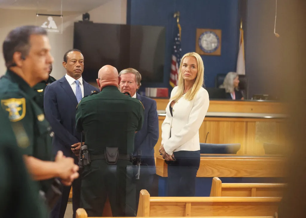 Tiger Woods in a suit and tie, standing next to a police officer, with his girlfriend Vanessa Trump looking on in court.