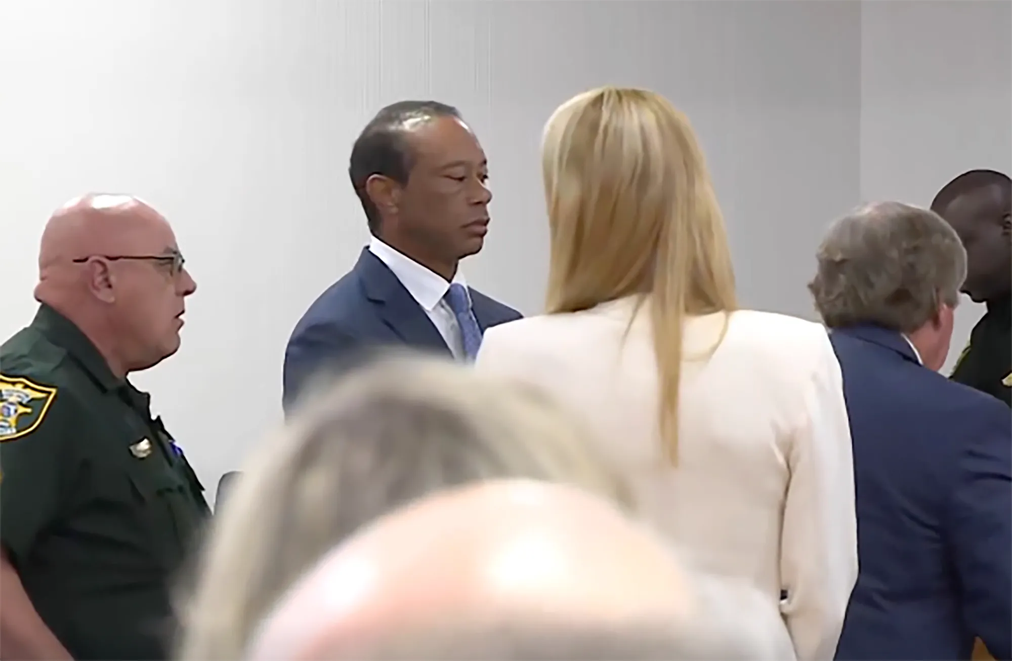 Tiger Woods in court, wearing a blue suit and light blue tie, with a blonde woman in a white blazer in the foreground.