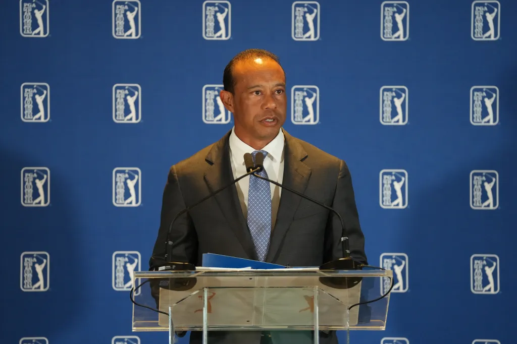 Tiger Woods speaking at a press conference with a PGA Tour logo backdrop.