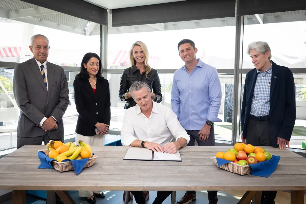 California Governor Gavin Newsom signs healthcare legislation, flanked by supporters behind a table with baskets of fresh fruit.
