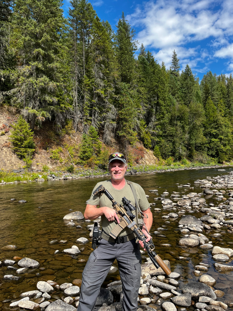 Todd Savage, a relocation consultant and former Marine, standing in a rocky river with a rifle and holster, smiling at the camera.
