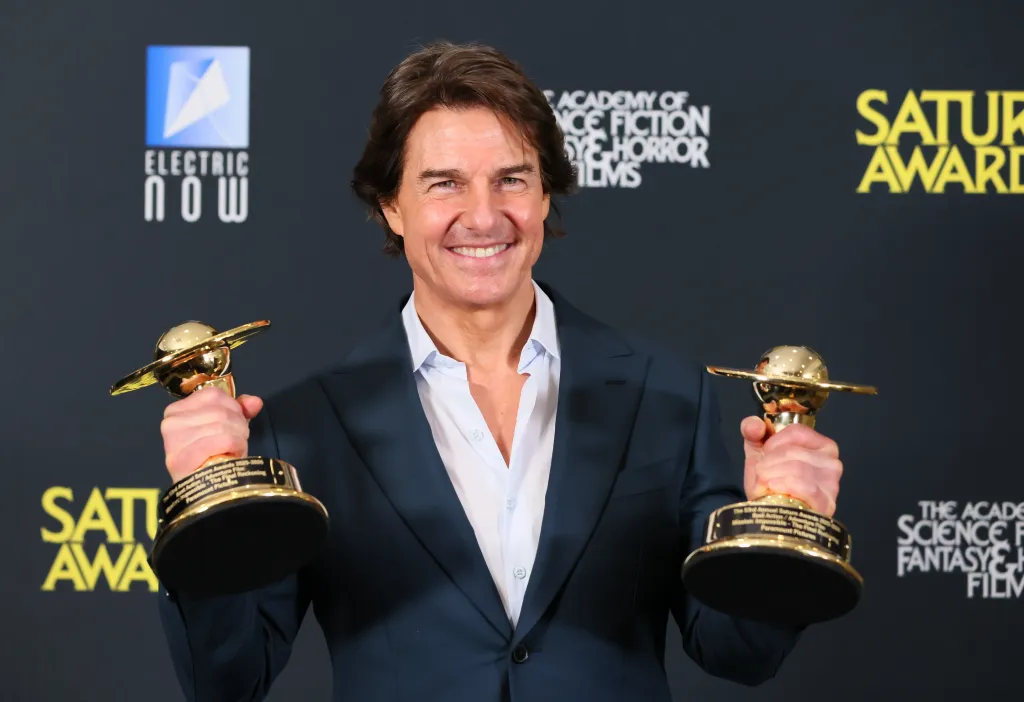 Tom Cruise, in a black suit and white shirt, holding two golden Saturn Awards.