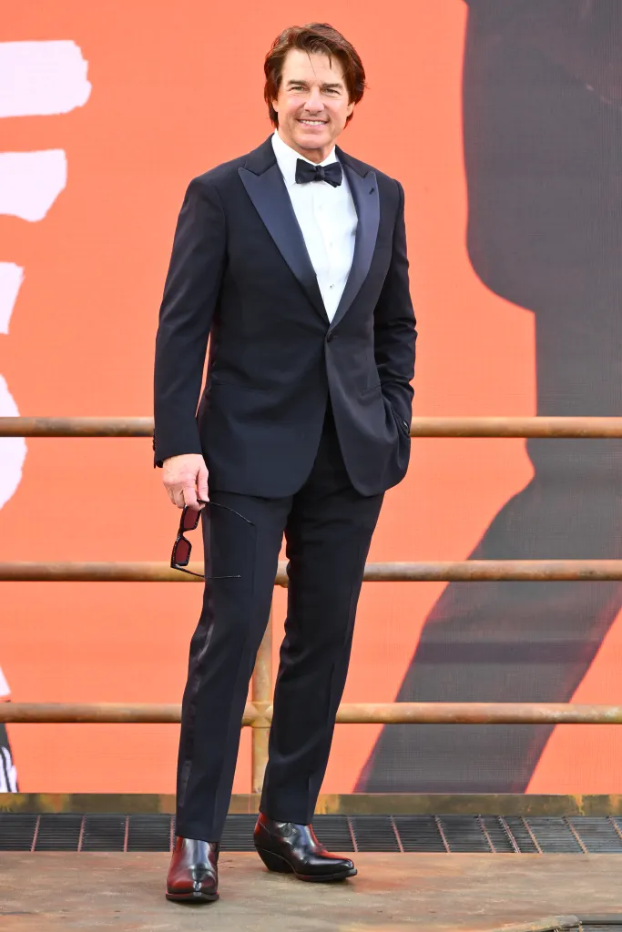 Tom Cruise in a black tuxedo with a white shirt and bow tie, holding sunglasses, on a red carpet.