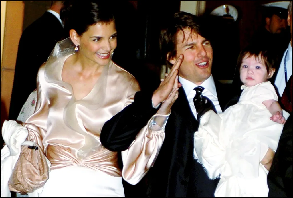 Katie Holmes,. in a cream silk shirt, smiling, Tom Cruise, in black suit, white shirt and black tie, holding their baby, Suri, in a white dress.