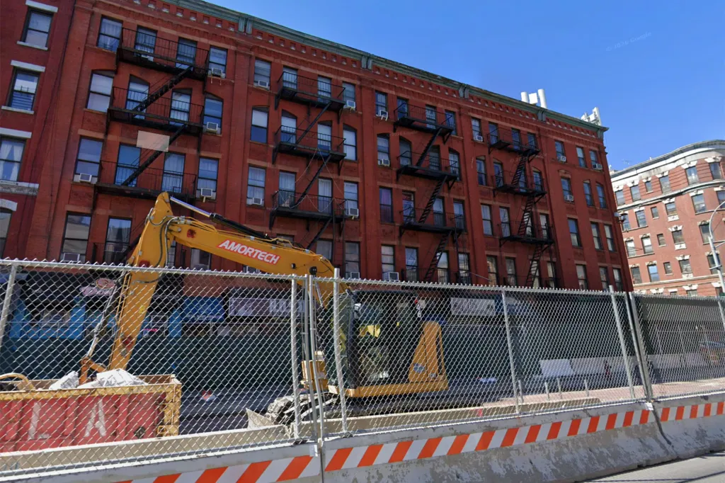 Construction site with an Amortech excavator in front of a red brick building with fire escapes in East Harlem.